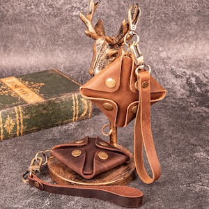 May include: Two square-shaped brown leather pouches with brass snap buttons and attached wrist straps. One pouch hangs from a metal hook, while the other rests on a circular wooden base. The leather features a matte finish and visible stitching.