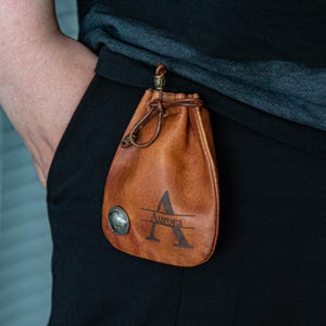 Puede incluir: Una bolsa de cuero marrón con cierre de cordón y un emblema metálico. La bolsa tiene el nombre "Aurora" y la letra "A" impresa. La bolsa está sujeta a una prenda negra.