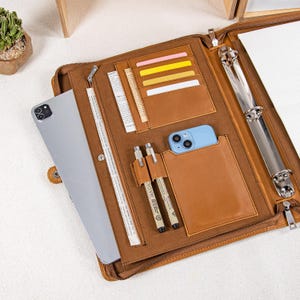 May include: A brown leather portfolio, open to reveal various compartments. It includes card slots, pen holders, and a smartphone pocket. An iPad is visible on the left. The portfolio has a three-ring binder and a zip closure.