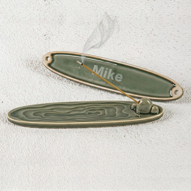 May include: Two green ceramic incense holders. One is oval with the name "Mike" etched on it, and the other has a wood grain design. An incense stick is lit and smoking in the top holder. The holders are on a white surface.