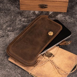 May include: A brown leather phone case with a brass button closure. The case is open, revealing a black smartphone. A small, brown leather key fob and a brown paper envelope tied with twine are also visible.