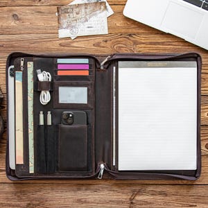 May include: Open brown leather portfolio with multiple pockets for a tablet, cards, pens, and a smartphone. A notepad is visible on the right. The portfolio is on a wooden surface, with a laptop and postcards in the background.