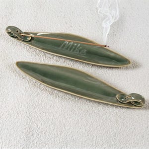 以下が含まれることがあります： 葉のようなデザインの、2つの緑色のセラミック香立て。1つは燃えているお香を持っており、煙が出ています。「Mike」という文字がホルダーに刻まれています。ホルダーは、質感のあるオフホワイトの表面にあります。