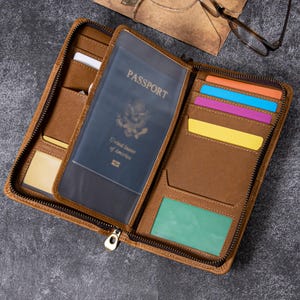 Puede incluir: Cartera de pasaporte de cuero marrón abierta con cierre de cremallera. La cartera está abierta, revelando un pasaporte estadounidense y varias ranuras para tarjetas de colores. La cartera está sobre una superficie gris oscuro.