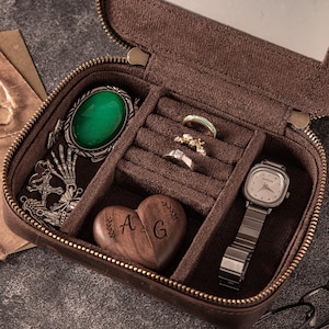 May include: Open brown leather jewelry case with a zip, containing a green gemstone pendant, rings, a watch, and a wooden heart with the letters "A G". The interior is lined with brown fabric.