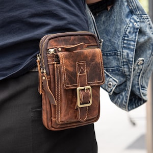 May include: A brown leather fanny pack with a distressed finish, featuring a front flap with a buckle closure and a zippered main compartment. The bag is worn on the waist and has a small pocket on the front. The bag is made of leather.