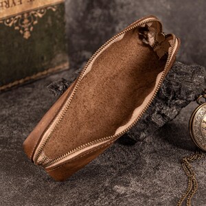 May include: Open brown leather pencil case with a brass zipper. The interior is a lighter shade of brown. A vintage pocket watch and a book are visible in the background. The pencil case is resting on a dark, textured surface.
