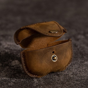 Puede incluir: Estuche de cuero marrón abierto con cierre de botón a presión. El estuche está diseñado para guardar objetos pequeños. El cuero tiene un aspecto envejecido, con grano visible y variaciones de color.