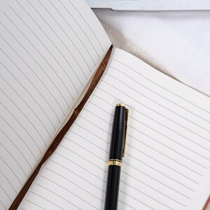 May include: An open notebook with lined pages and a black pen with gold accents. The notebook has a brown leather spine. The pen is positioned on the right side of the open notebook.