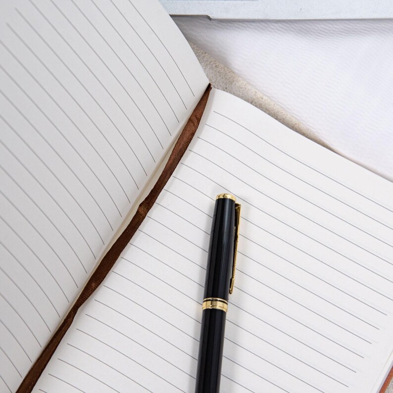 May include: An open notebook with lined pages and a black pen with gold accents. The notebook has a brown leather spine. The pen is positioned on the right side of the open notebook.