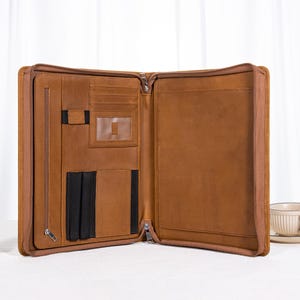 Op de afbeelding: Open, bruin leren portfolio met meerdere compartimenten en een lange ritssluiting. De binnenkant heeft kaartsleuven, penhouders en een doorzichtig ID-venster. De portfolio is ontworpen om documenten en andere benodigdheden in op te bergen.