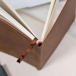 May include: A brown leather-bound journal with a textured cover and a brown ribbon bookmark. The pages are white and the binding is visible. The journal is open, displaying the interior pages.