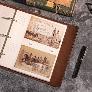 May include: A brown leather photo album with two vintage postcards inside. One postcard is labelled "CARTE POSTALE" and the other has a stamp that says "WE HAVE AT HELLO". A black pen with gold accents rests on the side.