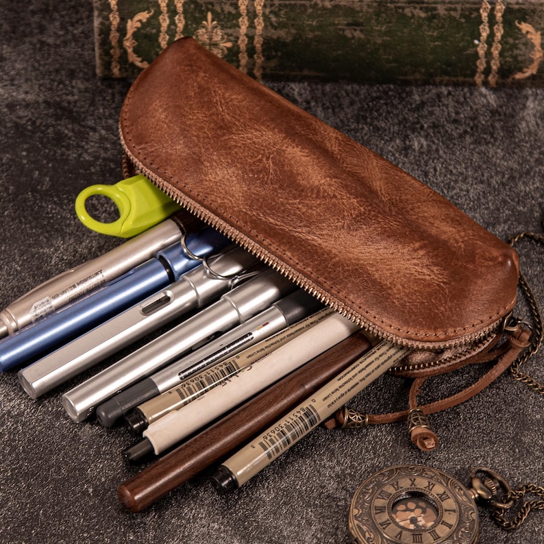 May include: A brown leather pencil case with a zipper, filled with various pens and pencils. The case is open, revealing the writing instruments inside. A vintage pocket watch with a chain is in the foreground.
