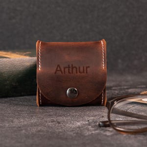 Op de afbeelding: Een klein, vierkant bruin lederen tasje met een klepsluiting die wordt vastgezet met een zilverkleurige metalen drukknoop. De naam Arthur is in het midden van de klep gedrukt. Het tasje is afgewerkt met opvallende witte stiksels.