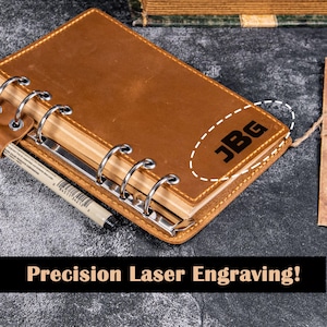 Op de afbeelding: Een bruin leren notitieboek met zilveren metalen ringen en de initialen "JBG" gegraveerd op de kaft. Een pen ligt op de open pagina's. De tekst "Precision Laser Engraving!" staat onderaan.