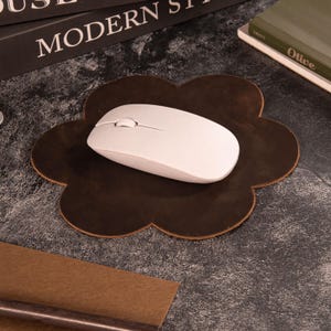 May include: A brown, flower-shaped leather mousepad with a white wireless mouse. The mousepad has a dark brown color and a unique, decorative design. The mouse is white with a scroll wheel and two buttons. The items are on a dark surface.
