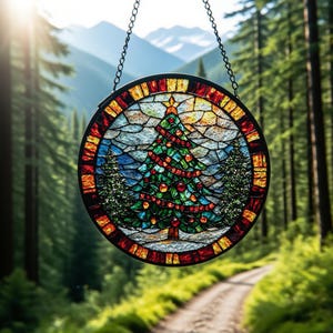 Op de afbeelding: Ronde gebrandschilderde glazen kerstboomversiering met een zwart metalen frame en ophangketting. De boom is groen met rode ballen en een gele ster. De achtergrond toont een bos en bergen.