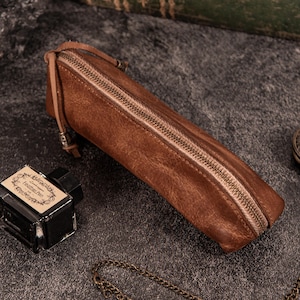May include: A brown leather pencil case with a zipper closure. The rectangular case is made of textured leather and has a small leather loop attached to the zipper pull. An ink bottle and chain are in the background.