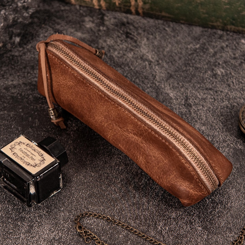 May include: A brown leather pencil case with a zipper closure. The rectangular case is made of textured leather and has a small leather loop attached to the zipper pull. An ink bottle and chain are in the background.