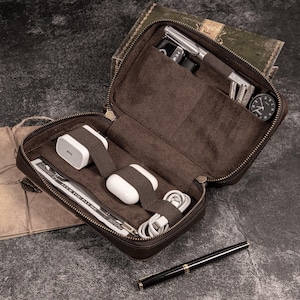Könnte beinhalten: Geöffnete braune Leder-Reise-Organizer mit Reißverschluss, enthält eine Uhr, Rasierer, Schlüssel, Ladegerät, Ohrhörer und einen Stift. Der Organizer ist offen und zeigt mehrere Fächer für Zubehör. Der Hintergrund ist eine dunkle, strukturierte Oberfläche.