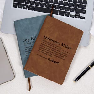 May include: Two leather-bound journals, one blue and one brown, are displayed on a laptop keyboard. The brown journal reads "Difference Maker" with a definition and the name "Robert." The blue journal says "Joy Bri."