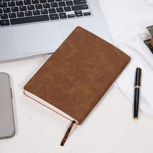 May include: A brown leather-bound notebook with a ribbon bookmark lies open on a white surface. A black pen with gold accents rests nearby. A laptop and a smartphone are partially visible in the background.