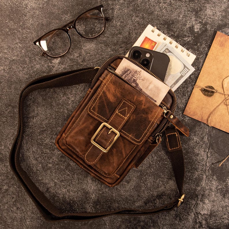 May include: A brown leather crossbody bag with a buckle closure, a front pocket, and a long adjustable strap. The bag is open, revealing a smartphone, cash, and a small notepad. A pair of eyeglasses rests nearby.