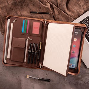 Puede incluir: Un portafolio de cuero marrón con cremallera, abierto para revelar un bloc de notas, una tableta y portalápices. El portafolio contiene bolígrafos, tarjetas y una regla. También se ve un bolígrafo negro y una herramienta con rueda.