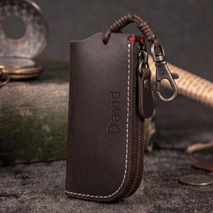 May include: A dark brown leather key holder with a zipper closure and a woven leather loop. The word "David" is embossed on the front. A silver-colored clasp is attached to the loop.