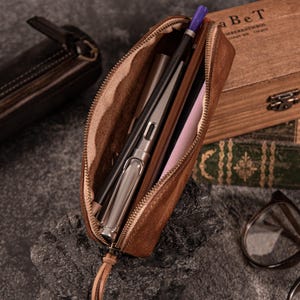 May include: A brown leather pencil case, partially unzipped, revealing pens and pencils inside. The case has a zipper closure and a leather pull. The interior is lined with a lighter-colored fabric. A second, closed pencil case is visible in the background.