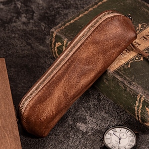 May include: A brown leather pencil case with a zipper closure. The case has a distressed texture and is rectangular in shape. The zipper is a lighter shade of brown. The case is lying on a dark surface next to a vintage book and a watch.