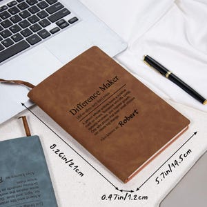 May include: A brown leather journal with the text "Difference Maker" and the name "Robert". The journal is open, showing the pages, and has a ribbon bookmark. Dimensions are 8.26 inches by 5.7 inches by 0.47 inches.