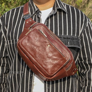 May include: A brown leather fanny pack with multiple zippered compartments and a shoulder strap. The bag has a rectangular shape with a front pocket and a metal buckle. The leather has a rich, textured appearance.