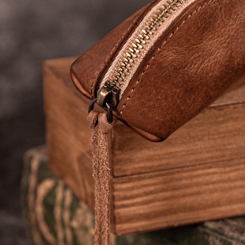 May include: Close-up of a brown leather pouch with a brass zipper and a leather tassel. The pouch is resting on a wooden surface, showcasing the texture of the leather and the details of the zipper.