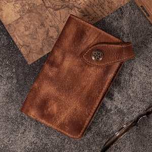 Puede incluir: Una cartera de cuero marrón con cierre de botón. La cartera tiene una superficie texturizada y una correa que se asegura con un botón decorativo. La cartera es rectangular y parece estar hecha de cuero de alta calidad.