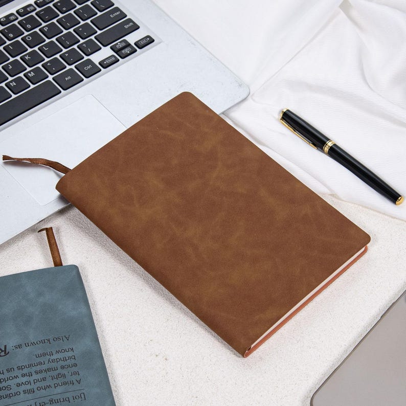 May include: A brown leather-bound notebook with a ribbon bookmark lies on a white surface next to a laptop and a black pen. The notebook has a textured appearance and a contrasting light brown edge.