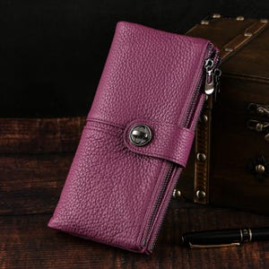 Puede incluir: Una cartera de cuero color ciruela con una superficie texturizada. La cartera presenta una correa envolvente con un cierre de metal y un cierre de cremallera. La cartera es rectangular y tiene un fondo oscuro.