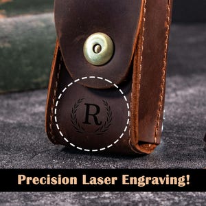 Könnte beinhalten: Nahaufnahme eines braunen Lederetuis mit einem Metallschnappverschluss. Das Etui hat ein kreisförmiges, eingraviertes Design mit dem Buchstaben "R" in einem Lorbeerkranz. Der Text "Precision Laser Engraving!" wird unten angezeigt.