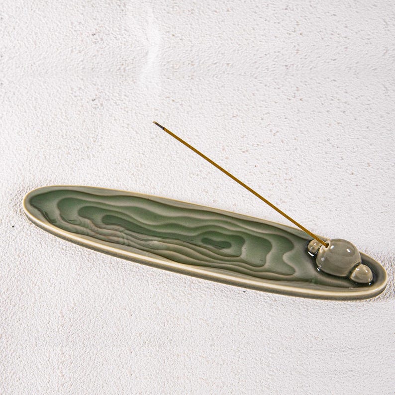 May include: A green ceramic incense holder with a layered design. The holder is oval-shaped with a small, round incense stick holder at one end. An incense stick is lit, with smoke rising.