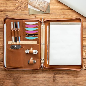 May include: Open brown leather portfolio with a zipper closure, containing a notepad, pen holders, card slots, and pockets for a phone and accessories. The portfolio is open on a wooden surface.