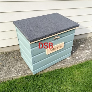 May include: A light blue wooden parcel delivery box with a dark grey textured lid. The front features a rectangular wooden plaque with the word PARCELS engraved in black, and the letters DSB are printed in red on the front panel.