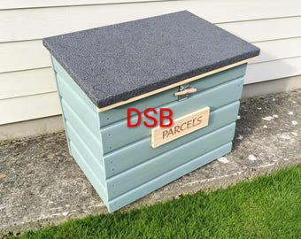 Può includere: Una cassetta per pacchi in legno azzurro con un coperchio grigio scuro testurizzato. La parte anteriore presenta una targa rettangolare in legno con la parola PARCELS incisa in nero e le lettere DSB stampate in rosso sul pannello.
