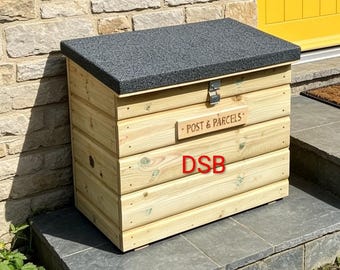 Può includere: Una scatola rettangolare in legno con coperchio testurizzato grigio scuro e chiusura in metallo. Il pannello frontale presenta una targa orizzontale in legno con la scritta "POST & PARCELS" e le lettere rosse "DSB" sul legno chiaro.