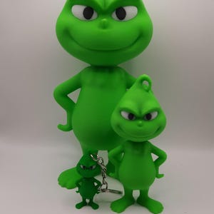 Könnte beinhalten: Eine Sammlung grüner Grinch-Figuren in verschiedenen Größen. Die größte Figur steht aufrecht mit verschränkten Armen, während eine kleinere die Pose nachahmt. Ein Grinch in Schlüsselanhängergröße vervollständigt das Set, alle mit dem charakteristischen Grinsen der Figur.