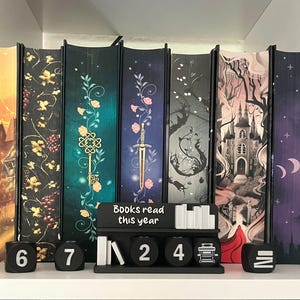 Könnte beinhalten: Ein dekoratives Bücherregal-Trennwand-Set mit Fantasy-Motiven. Das Set enthält einen Zähler mit dem Text "Books read this year" und Würfel, um die Anzahl der gelesenen Bücher zu verfolgen. Die Trennwände zeigen Illustrationen von Burgen, Schlüsseln und Schwertern.