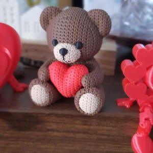 Könnte beinhalten: Handgefertigte braune Teddybär-Figur, die ein rotes Herz hält. Der Bär hat eine hellbraune Schnauze und Pfoten, mit schwarzen Augen und Nase. Der Bär sitzt auf einer Holzoberfläche. Das Bild ist unscharf.