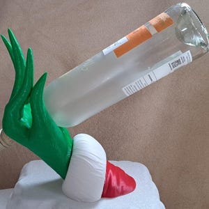 Könnte beinhalten: Ein grüner Flaschenhalter in Handform mit langen, schlanken Fingern, die eine klare Glasflasche halten. Die Basis des Halters ist mit einer weißen Manschette und einem roten Abschnitt verziert, die an einen Ärmel erinnern. Die Flasche hat ein weißes Etikett.