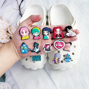 May include: A hand holding a collection of colorful cartoon character shoe charms. The charms feature various designs, including anime-style figures, a heart-shaped charm with "K POP" text, and a blue cat. White clogs are in the background.