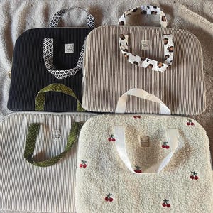 Peut inclure: Quatre housses pour ordinateur portable rectangulaires avec poignées. Une en velours côtelé noir à motifs floraux, une en velours côtelé beige à motif léopard, une en velours côtelé gris clair à pois verts, et une en sherpa crème brodée de cerises avec des poignées blanches.
